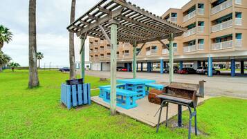 Un espace de pique-nique abrité, doté de bancs bleus et d’un barbecue, situé devant un immeuble à plusieurs étages avec des balcons.
