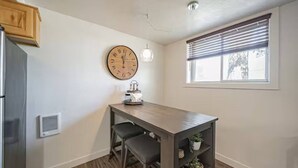 A modern kitchen with a wooden table, two bar stools, a clock, a pendant light, and a window with blinds. - Gate City Grotto (New Owners) (Pocatello)