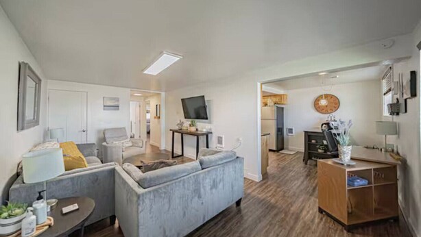 A modern living room with a grey sofa, a small table, and a TV. There's a kitchen area with a fridge and a clock on the wall. - Gate City Grotto (New Owners) (Pocatello)