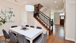 A dining area with a white table and grey chairs, a staircase with wooden steps and black railings, and a modern abstract painting on the wall. - Bright & Spacious 5BR Home Near Capitol Hill • Free Parking (Washington DC)