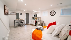 A modern kitchen with white cabinets, a microwave, and an oven. There is a dining area with a table and chairs. The living room has a white sofa with a red pillow and an orange throw blanket. - Comfy 1BR Basement DC | Sleeps 5 | Near Metro (Washington DC)
