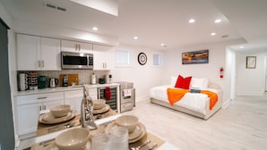 A modern kitchen with white cabinets, a microwave, and a coffee machine. There is a dining area with a table set for two, and a bed with white bedding and an orange throw blanket. - Comfy 1BR Basement DC | Sleeps 5 | Near Metro (Washington DC)