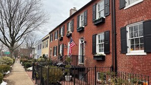 A row of brick townhouses with black shutters, a flag, and a small garden area. - Historic English Basement – Elegant Old Town, Alexandria Retreat (Alexandria)