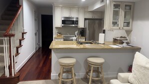 A modern kitchen with white cabinets, stainless steel appliances, and a granite countertop. There are two wooden stools and a white couch in the adjacent living area. - Historic English Basement – Elegant Old Town, Alexandria Retreat (Alexandria)