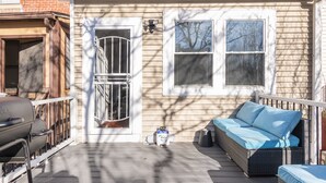 Eine überdachte Veranda mit Grill, einem Rattan-Sofa und einem Fenster mit Jalousien.