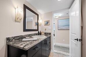 A bathroom with a granite countertop, a large mirror, and a sink. There are two framed pictures on the wall, a towel on the counter, and a door leading to another room. - 11th Floor Corner Unit - Views Galore! (Pensacola Beach)