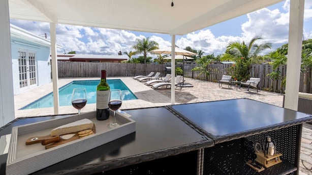 A poolside area with a covered seating area, a pool, and a view of a house in the background. - Sunshine Retreat! New! Sparkling Blue Pool And Spa (West Palm Beach)
