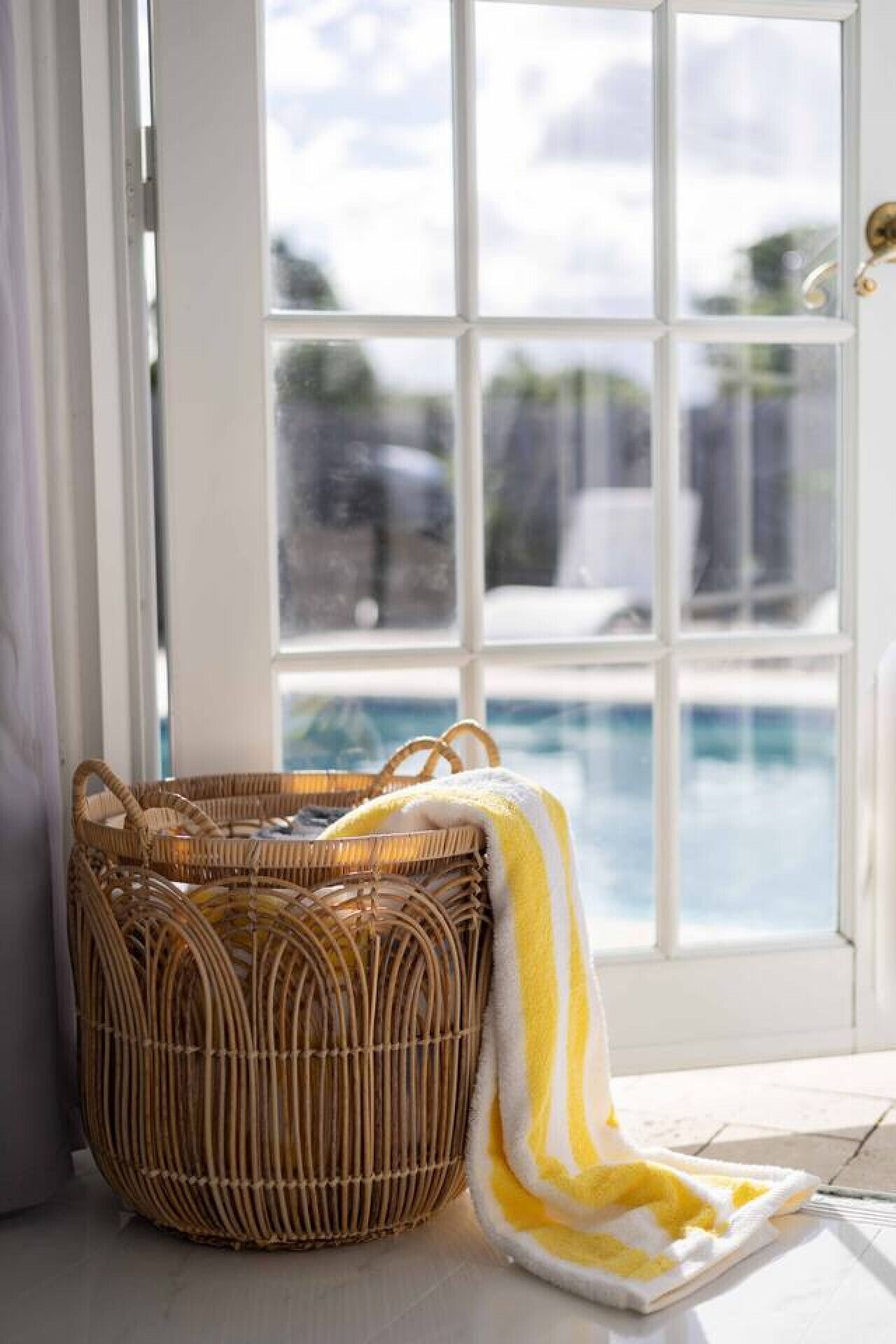 A wicker basket with a yellow and white striped towel beside a glass door leading to a pool area.