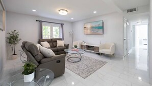 A modern living room with a reclining sofa, a glass coffee table, a white cabinet, and a painting on the wall. - Sunshine Retreat! New! Sparkling Blue Pool And Spa (West Palm Beach)