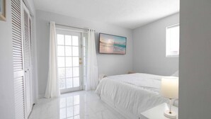 A bedroom with a bed, a nightstand, a lamp, a painting, and a door with curtains. - Sunshine Retreat! New! Sparkling Blue Pool And Spa (West Palm Beach)