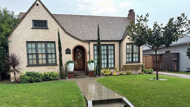A two-story house with a stone facade, a front entrance with a wooden door, and a small front yard with a concrete pathway leading to the door. - Charming Furnished Tudor Revival in the Heart of Historic California Heights (Long Beach)