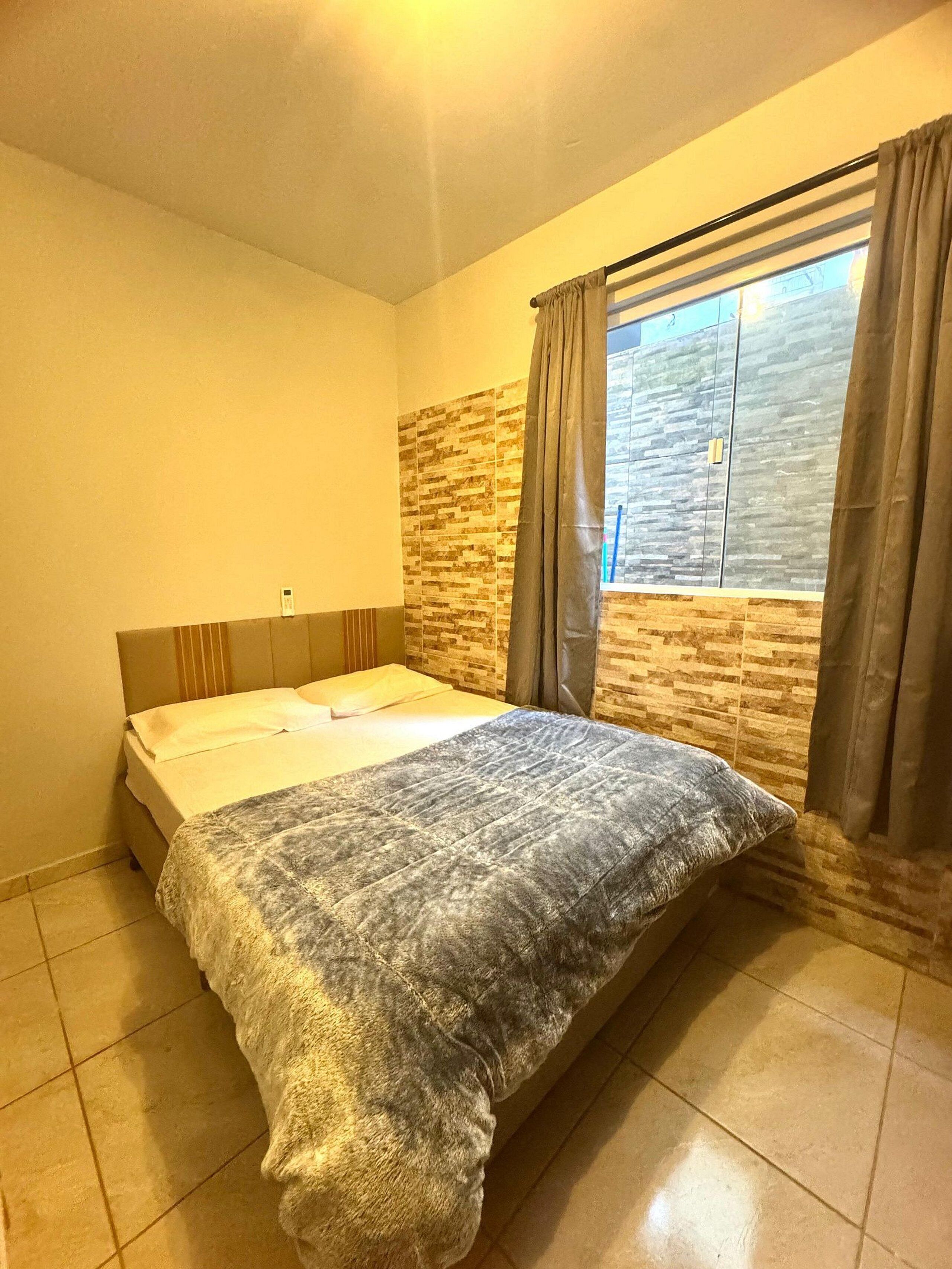 A bedroom with a bed, a window with curtains, a stone wall, and a tiled floor.