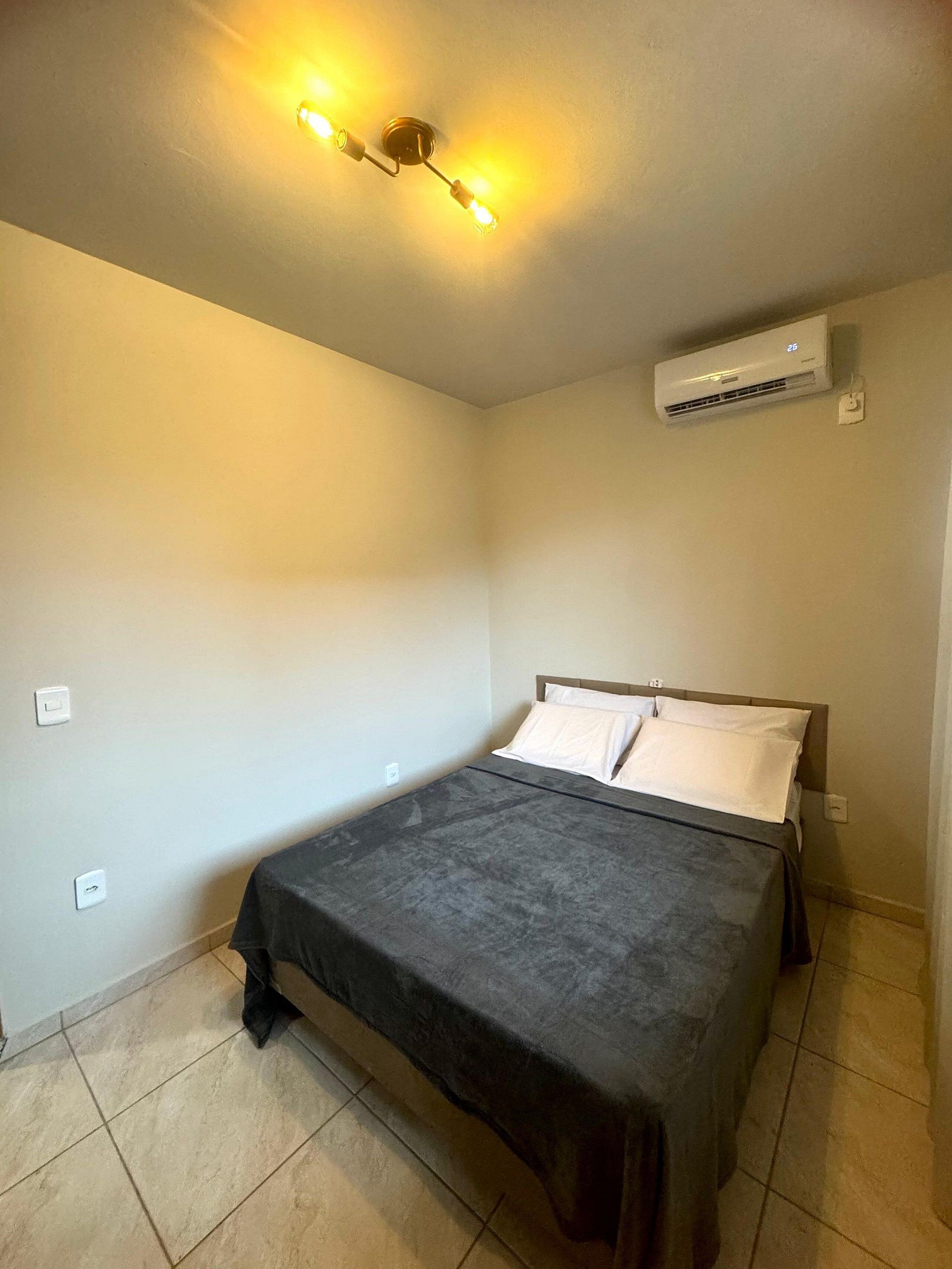 A small, clean room with a single bed, a ceiling light fixture, and an air conditioning unit.