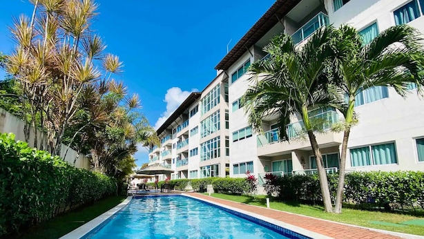 Une piscine à l’eau d’un bleu cristallin, entourée d’une végétation luxuriante et bordée d’un immeuble à plusieurs étages doté de balcons.