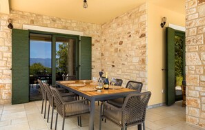 A dining area with a stone wall backdrop, a wooden table, wicker chairs, and a view of the outdoors. - Amazing home in Vrbnik with sauna (Vrbnik)