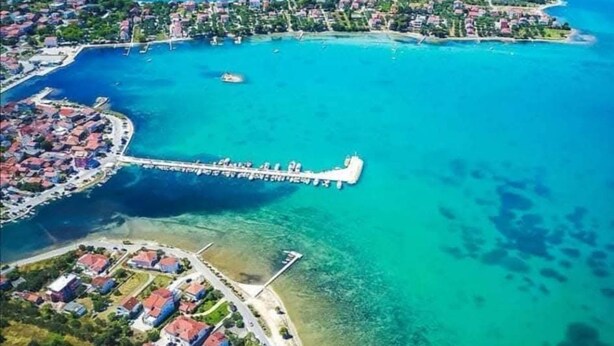 Vista aérea de uma área costeira com um píer branco proeminente que se estende para o mar azul-turquesa, cercado por edifícios residenciais e águas cristalinas.
