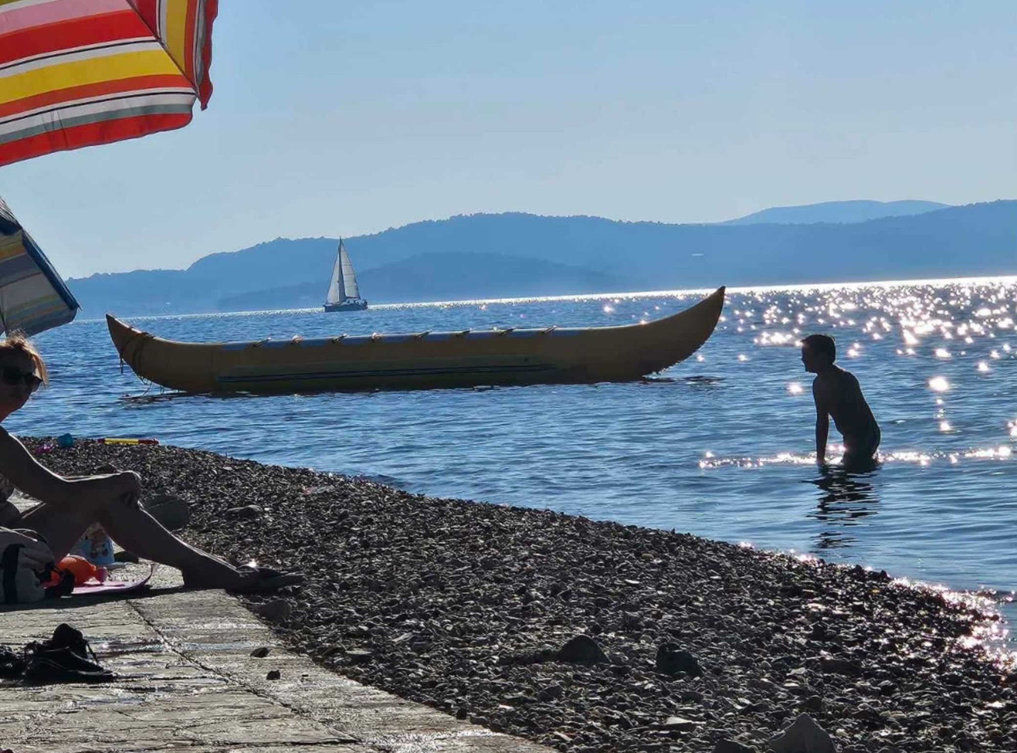 A rocky shoreline with a canoe and a sailboat in the water, and a person standing in the water.