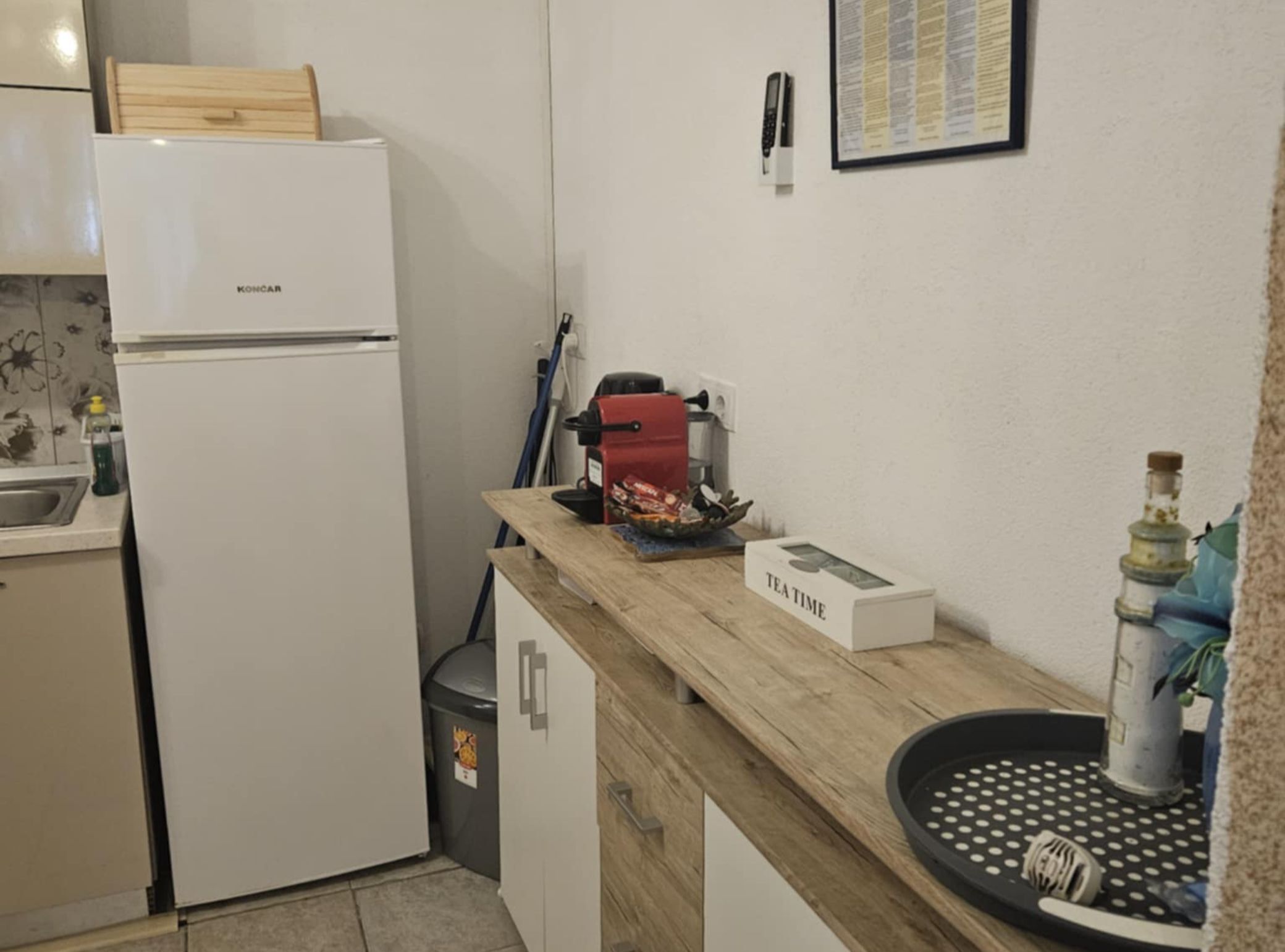 A compact kitchen with a white refrigerator, a coffee machine, a tea box, and a trash bin.