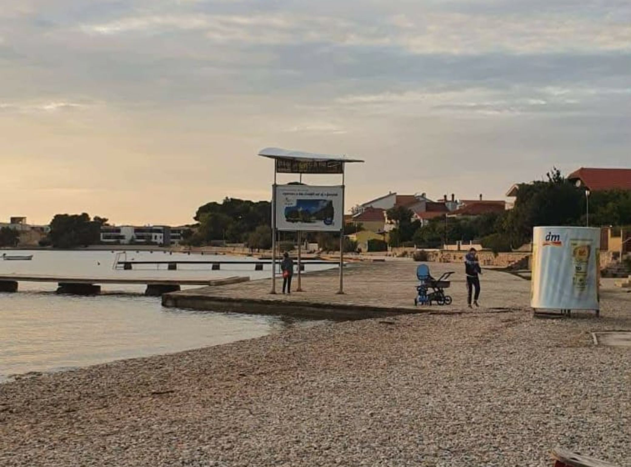 Ein Kiesstrand mit einem Schild, einem abgestellten Kinderwagen und einem Gewässer im Hintergrund.