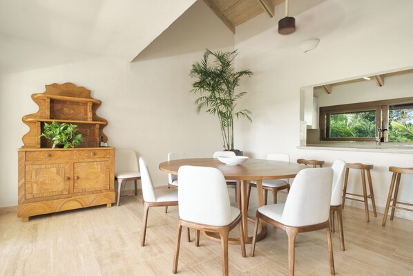 Een eetgedeelte met een ronde houten tafel, beklede witte stoelen, een houten kast met planten, en een groot raam met uitzicht op groen.