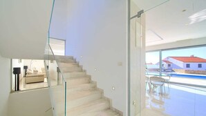 A modern interior with a glass staircase leading to a balcony with a view of houses and a swimming pool.
