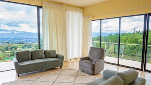 Amplia sala de estar con un sofá grande, un sofá más pequeño y una silla, todo sobre un piso de madera con alfombra estampada. La habitación cuenta con ventanas grandes que ofrecen vistas a un paisaje verde y montañas.