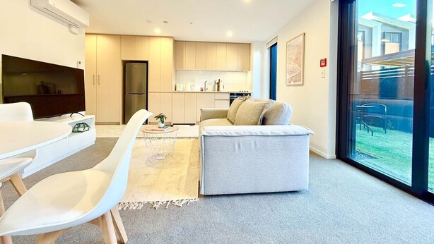 Ein modernes Wohnzimmer mit Sofa, einem Esstisch mit Stühlen, einer Küchenzeile und einem großen Fenster mit Blick ins Freie.