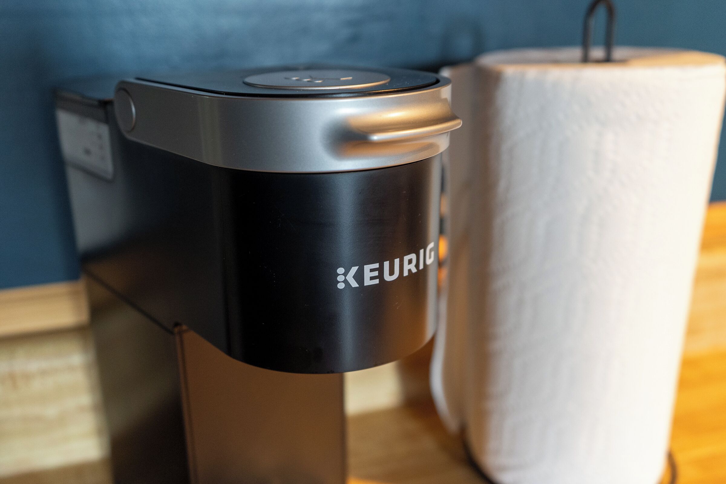 Una cafetera Keurig negra con un panel de control, un dispensador de toallas de papel blanco y una superficie de madera.