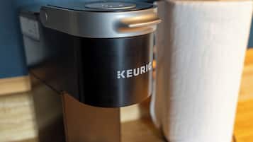 Una cafetera Keurig negra con un panel de control, un dispensador de toallas de papel blanco y una superficie de madera.