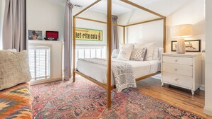 Une chambre à coucher avec un lit à baldaquin, un tapis à motifs, une table de chevet blanche et une fenêtre avec des stores.