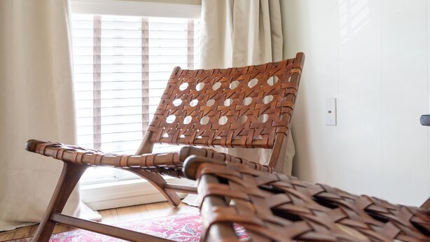 Une chaise en osier, dotée d’un dossier et de bras tressés, placée dans une pièce où se trouvent un tapis à motifs et une fenêtre avec des stores.