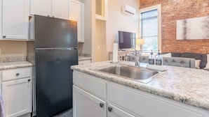 Una cocina con armarios blancos, un refrigerador negro, encimera de granito, un fregadero y una zona de comedor con sofá gris.