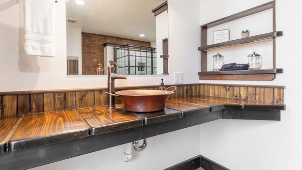 Un baño con tocador de madera, un lavamanos de cobre y un espejo encima. Hay una toalla colgada en la pared y una repisa con frascos y una planta.