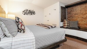 Un dormitorio con cama, un asiento junto a la ventana con almohadones, un reloj de pared y una pared de ladrillo.