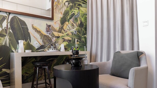 Een moderne woonkamer met een eethoek, met een grote tropische muurschildering, een ronde zwarte tafel en een grijze fauteuil.