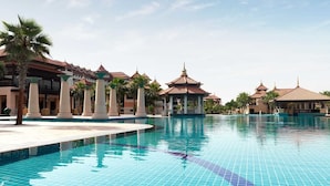 Una gran piscina con bordes de azulejos, rodeada de palmeras y edificios de estilo tradicional con techos de tejas rojas.