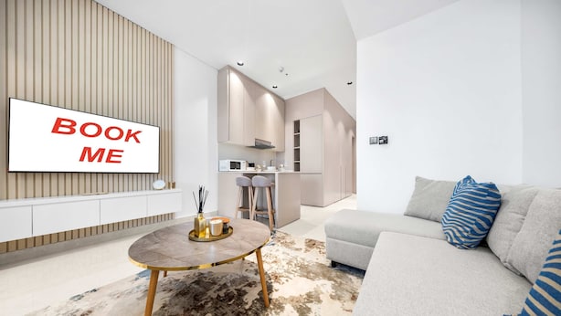 Un salón moderno con un sofá, una mesa de centro y un televisor que muestra “BOOK ME”.