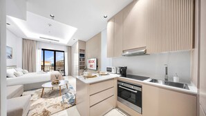 Cocina moderna con horno integrado, microondas y lavaplatos. Hay una zona de comedor con una mesa pequeña y sillas, además de un sofá. La habitación cuenta con un ventanal con vistas a un edificio.