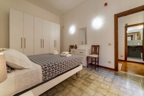 Ein Schlafzimmer mit Bett, Kommode, Stuhl und Spiegel, durch eine offene Tür ist ein Badezimmer sichtbar.