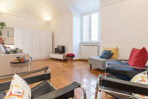 Ein Wohnzimmer mit Sofa, Sessel, Fernseher, Heizkörper und Fenster.