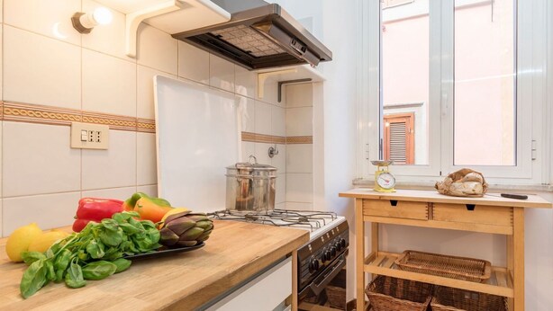 A kitchen with white tiles, a gas stove, a wooden table with baskets, and a window with blinds. - Casa dell'Arco - WelcometoRhome (Roma)