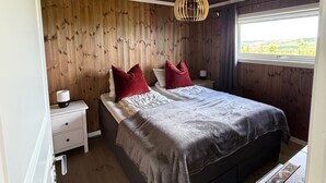 A bedroom with a wooden ceiling, a bed with a grey blanket, two red pillows, a white nightstand, and a window with a view. - Family Cabin Budor - 60 MIN From OSL - Sauna & Hedmarksvidda (Loten)