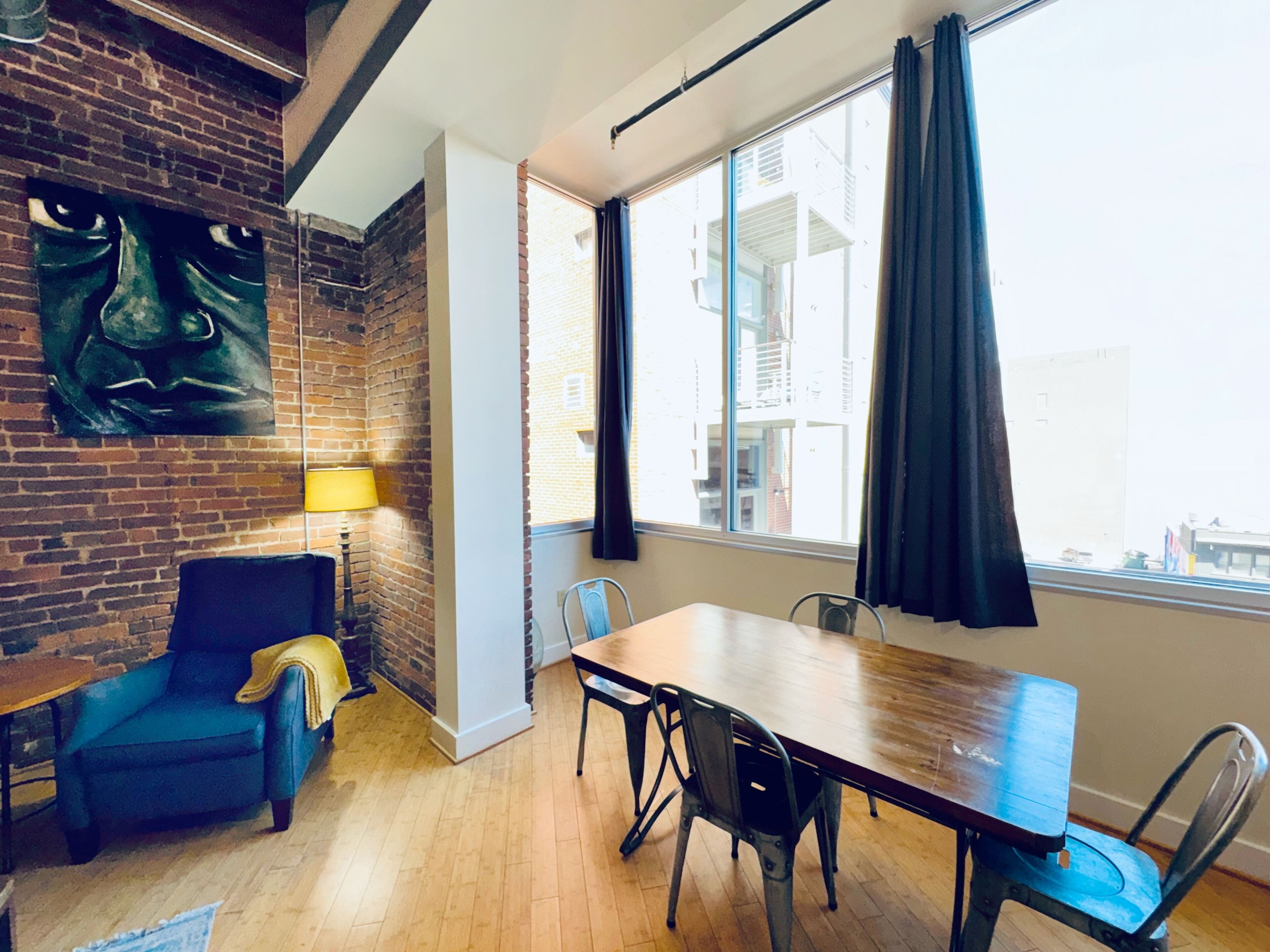 A modern room with a brick wall, a large window with curtains, a dining table with chairs, a blue armchair, and a lamp.