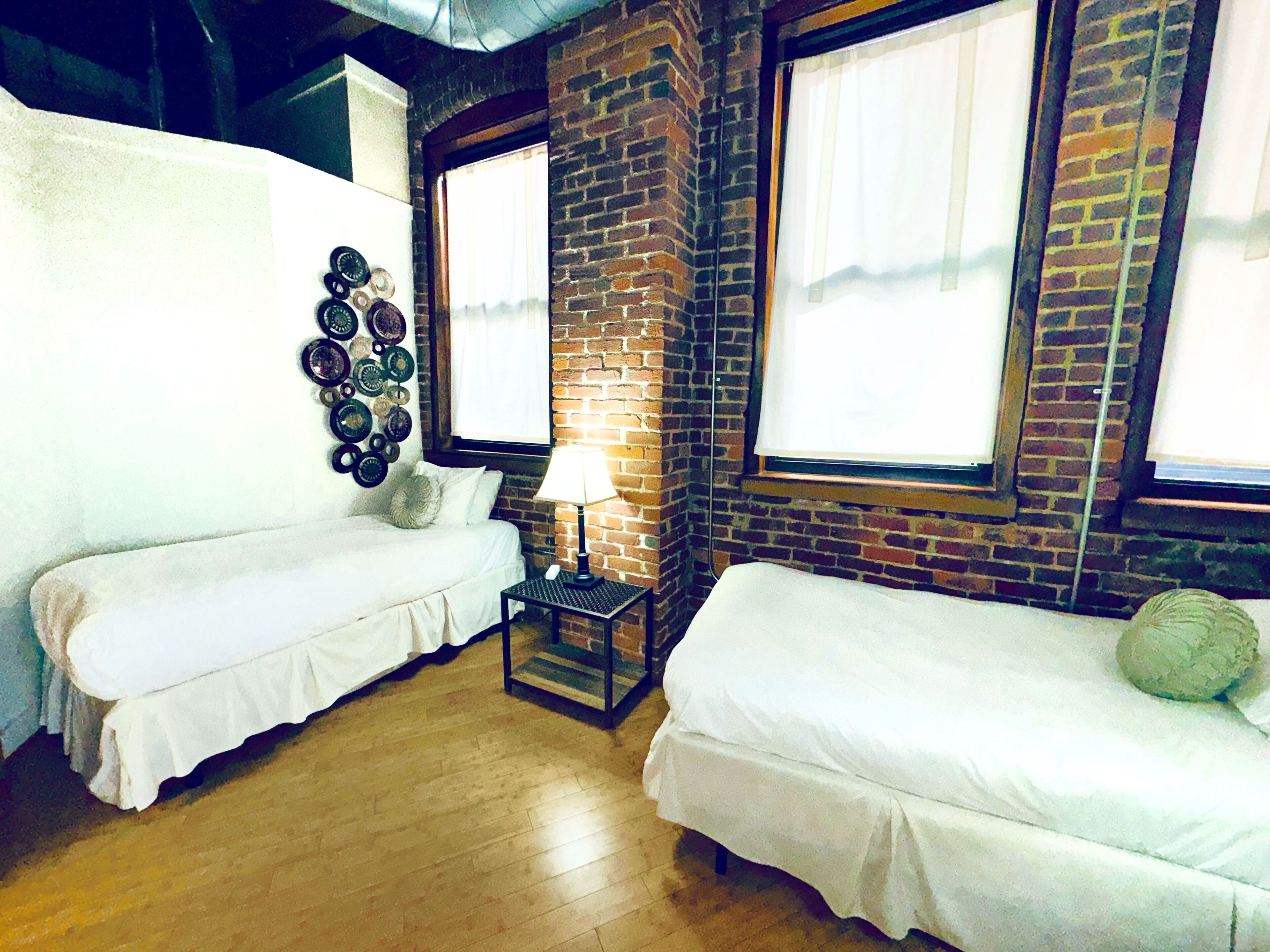 A room with two single beds, a small table with a lamp, a wall-mounted clock, and a brick wall.