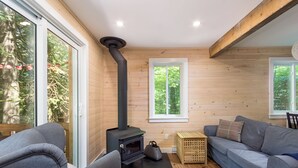 Una habitación acogedora con estufa de leña, sofás grises y amplios ventanales con vistas a la vegetación.