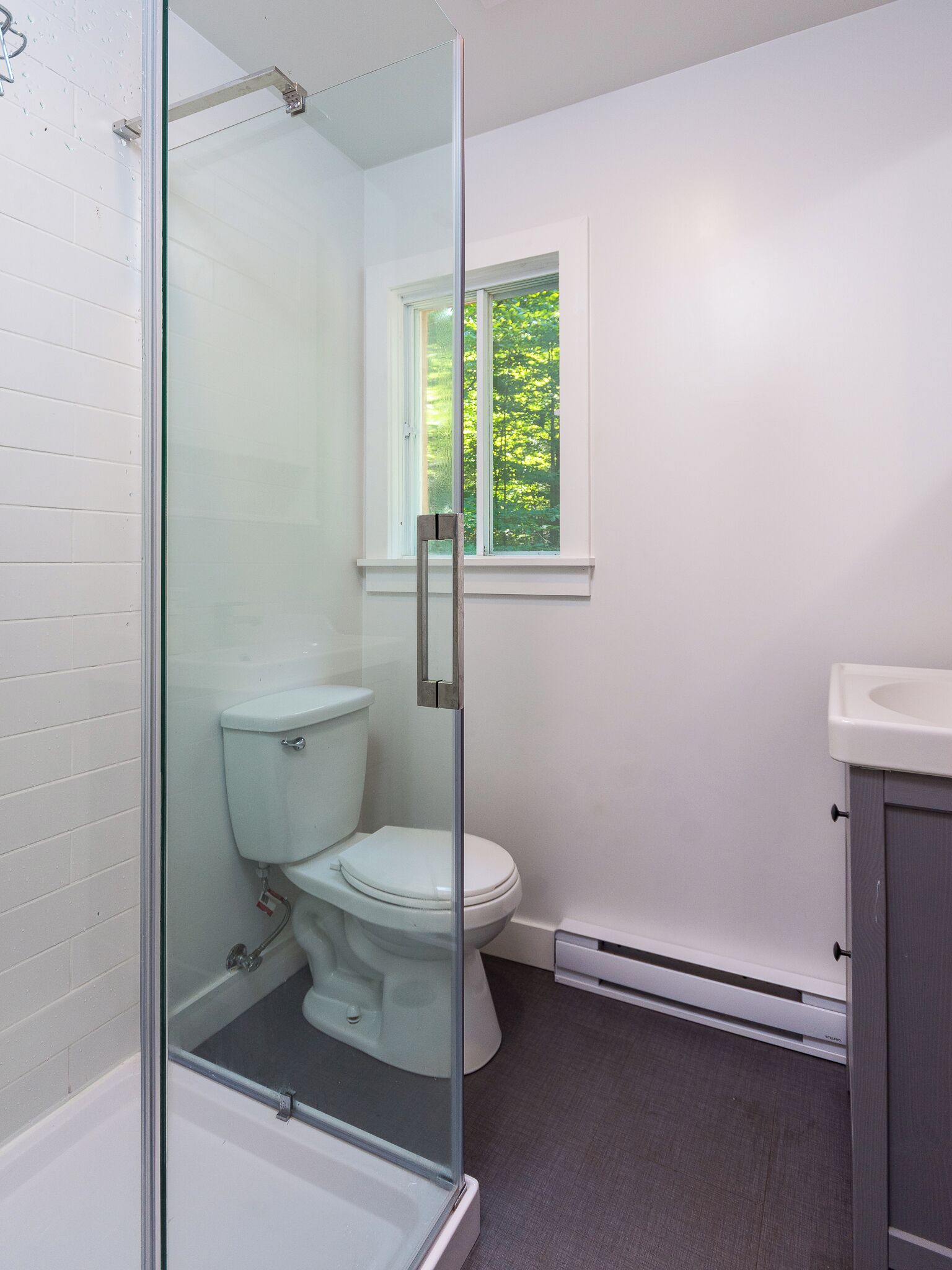 A bathroom with a glass-enclosed shower, a toilet, a window with a view of greenery, and a sink.