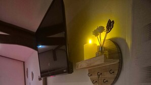 A hotel room with a bed, a red chair, a TV mounted on the wall, a bedside table with a remote control, and a wall-mounted shelf with decorative items. - Romantic room (Les Arcs)