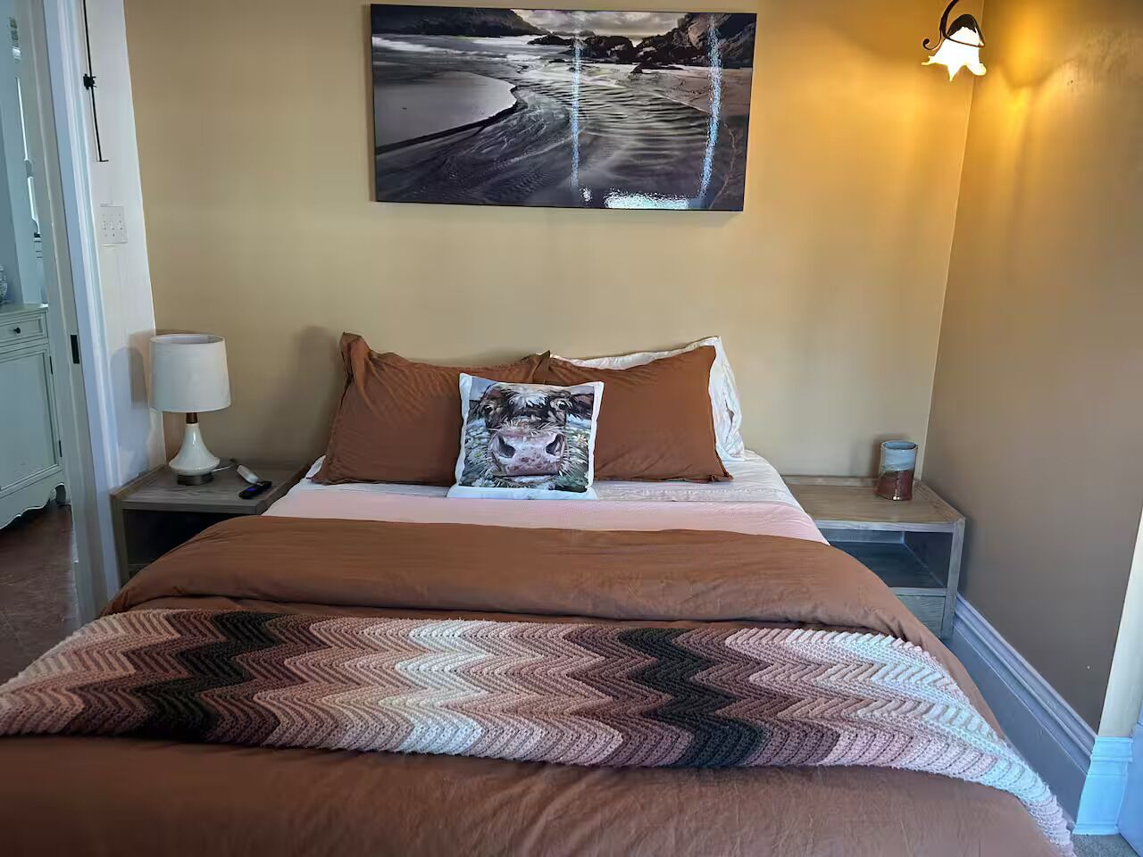 Een net opgemaakt bed met bruin beddengoed en een decoratief kussen, een aan de muur hangend schilderij en een bedlampje.