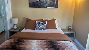 Een net opgemaakt bed met bruin beddengoed en een decoratief kussen, een aan de muur hangend schilderij en een bedlampje.