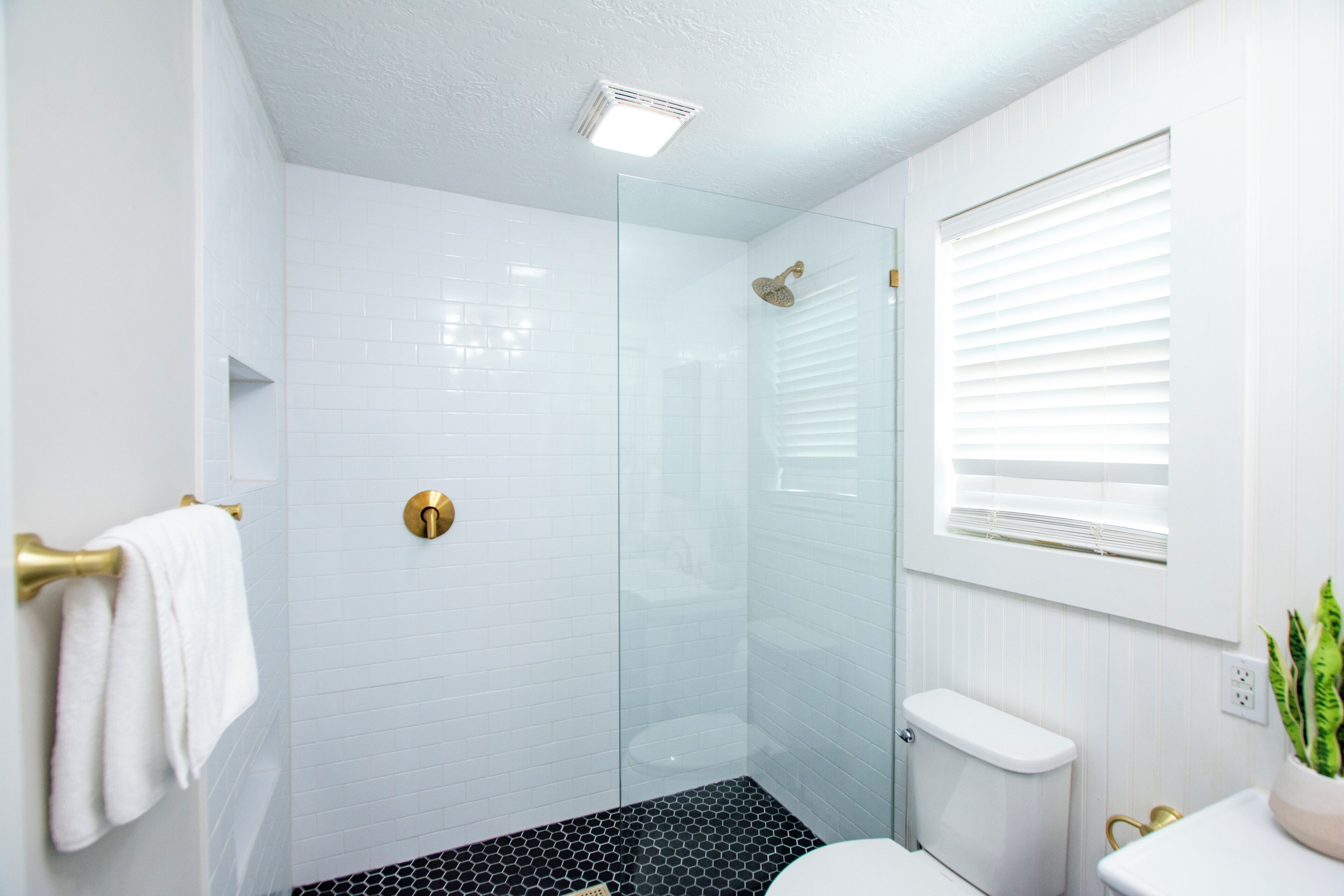 A modern bathroom with white tiles, a glass shower enclosure, a toilet, and a window with blinds.